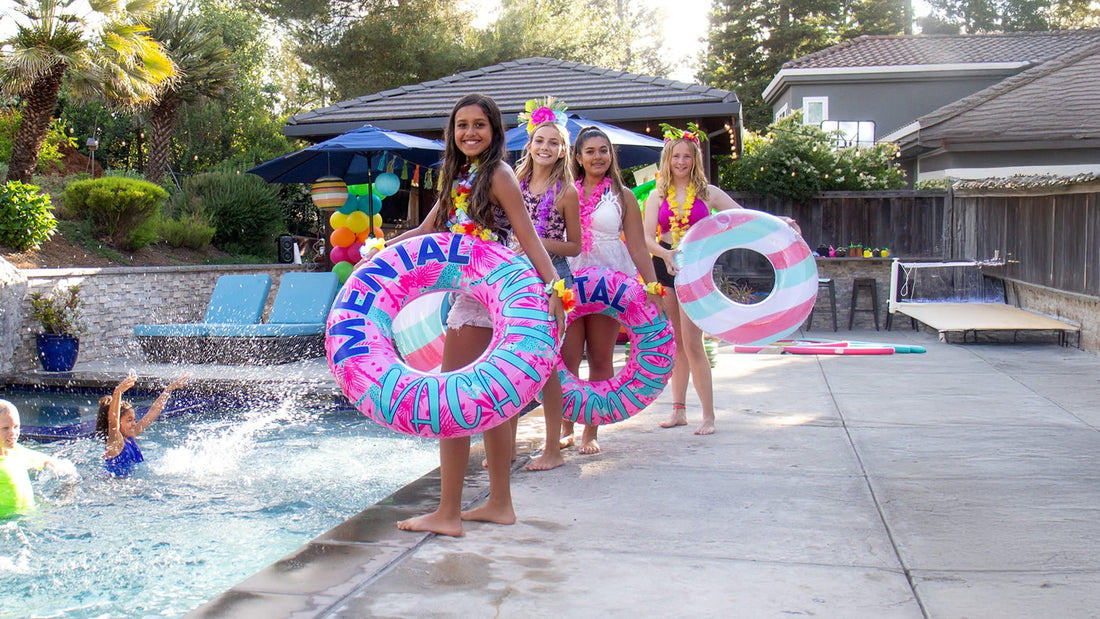 Four girls in floral leis and swimwear pose poolside with colorful inner tubes during a backyard summer party.