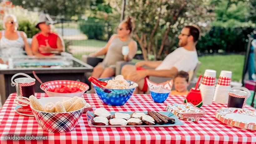 Outdoor gathering with people sitting around a table set with food and drinks.
