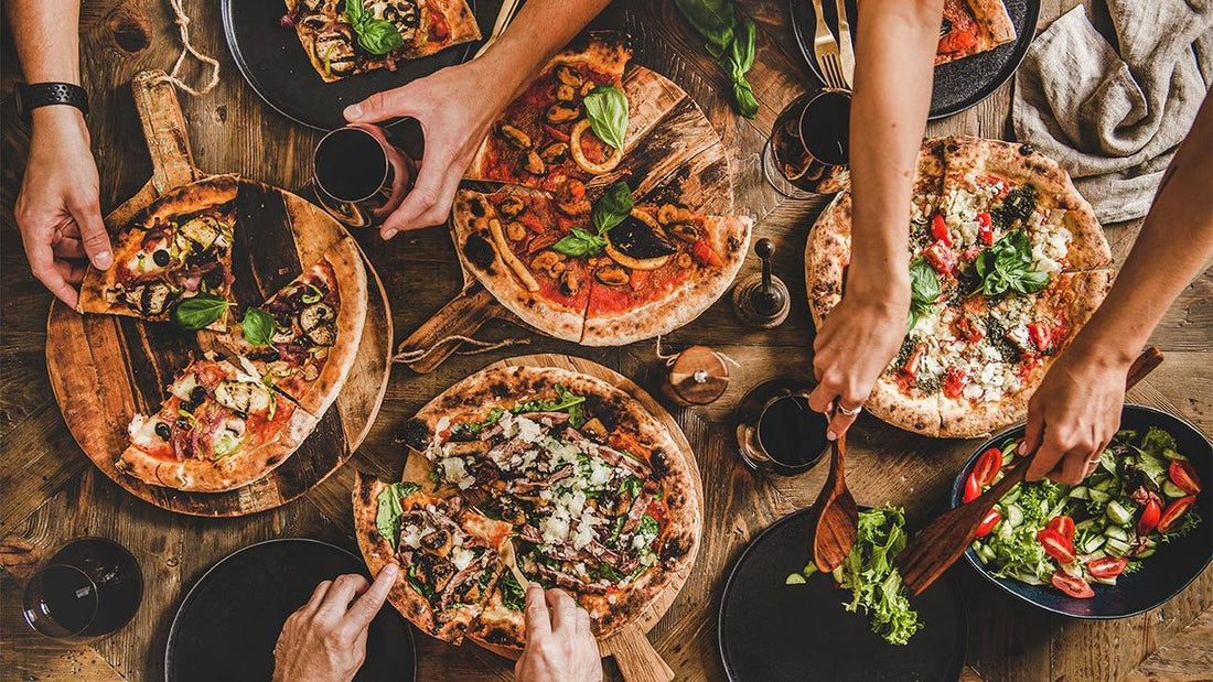 An aerial view of people sharing several artisan pizzas and a fresh garden salad on a rustic wooden table.