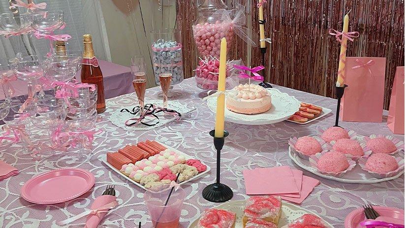 A festive dessert table decorated in pink and white features an assortment of sweets, champagne glasses, and lit candles arranged on a lace tablecloth.