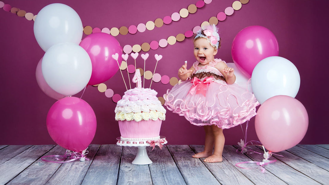 A joyful baby girl in a pink tutu stands on a wooden floor next to a large cupcake-shaped cake and balloons against a purple backdrop.