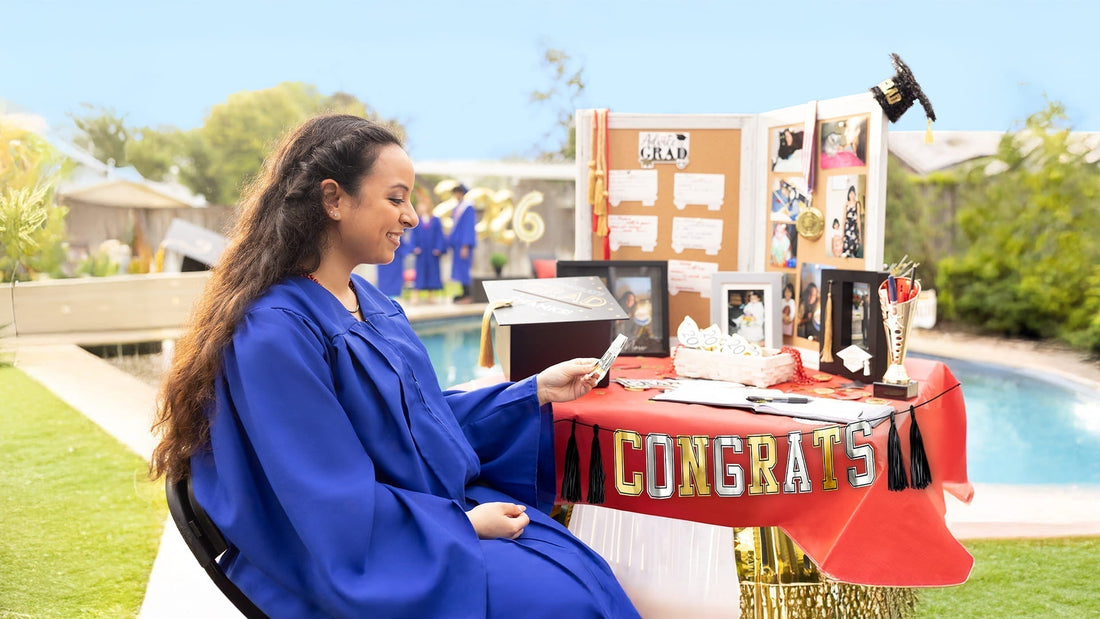 A smiling graduate in a blue gown sits at a decorated outdoor display table featuring photos, a guest book, and a "Congrats" banner.