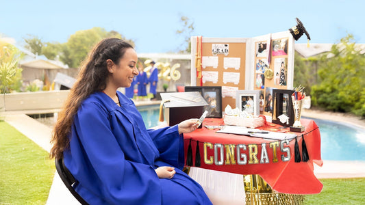 A smiling graduate in a blue gown sits at a decorated outdoor display table featuring photos, a guest book, and a "Congrats" banner.