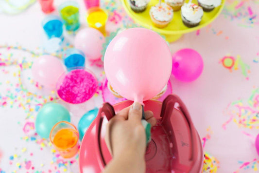 Hand inflating a pink balloon with a helium tank on a colorful party table with balloons, confetti, and cupcakes.