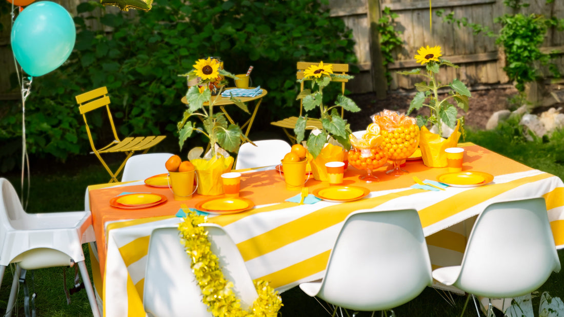 Outdoor party table set with orange and yellow decorations, sunflower centerpieces, and floating balloons against a garden backdrop.