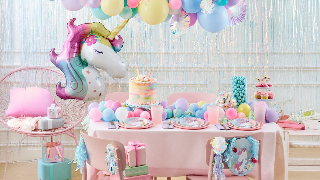 A pastel-colored unicorn-themed party table features a colorful cake, unicorn plates, balloons, and gifts against a shimmering fringe backdrop.