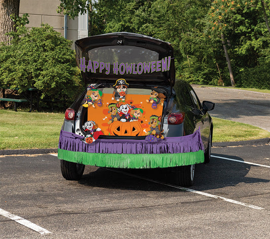A car trunk decorated for Halloween, with a purple and green fringe banner, a large orange jack-o'-lantern, cartoon characters, and a 'HAPPY HALLOWEEN' sign on the open trunk, parked in a paved outdoor area.