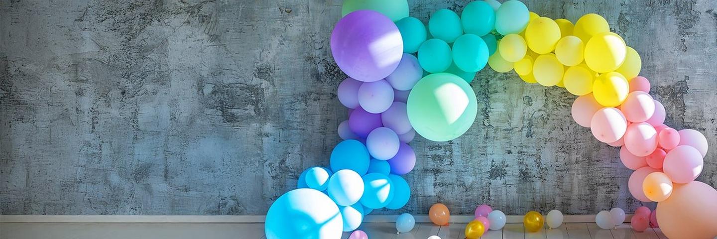 A vibrant, multi-colored balloon arch in a rainbow gradient spans across a rustic, grey concrete wall.