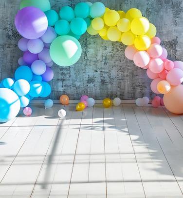 A vibrant, multi-colored balloon arch in a rainbow gradient spans across a rustic, grey concrete wall.