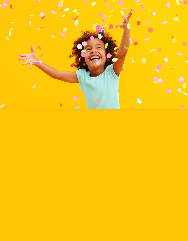 A joyful young child with curly hair and a wide smile stands against a bright yellow background with their arms outstretched as colorful pastel confetti falls around them.