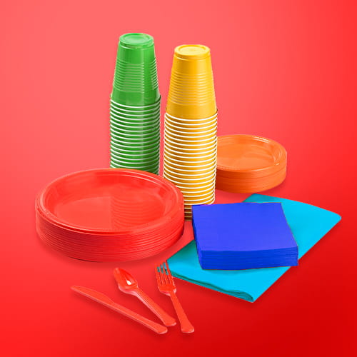 A colorful assortment of disposable party tableware, including stacks of red and orange plates, green and yellow cups, blue napkins, and red plastic cutlery against a red background.
