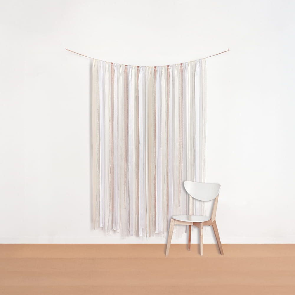 A minimalist photo booth backdrop featuring vertical ribbons of white, cream, and blush pink hanging against a white wall next to a modern white chair.