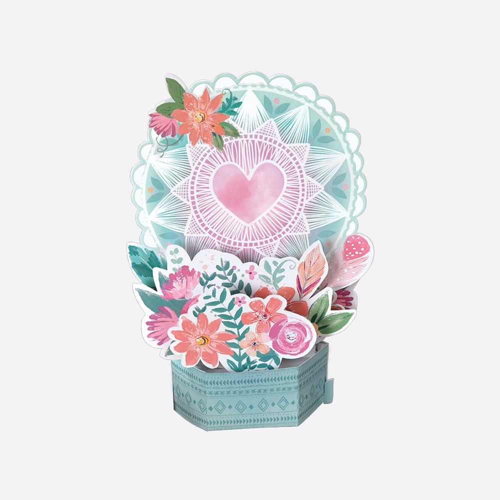 A colorful boho-style 3D pop-up tabletop decoration featuring a pink heart inside a teal mandala, accented by floral and feather cutouts.