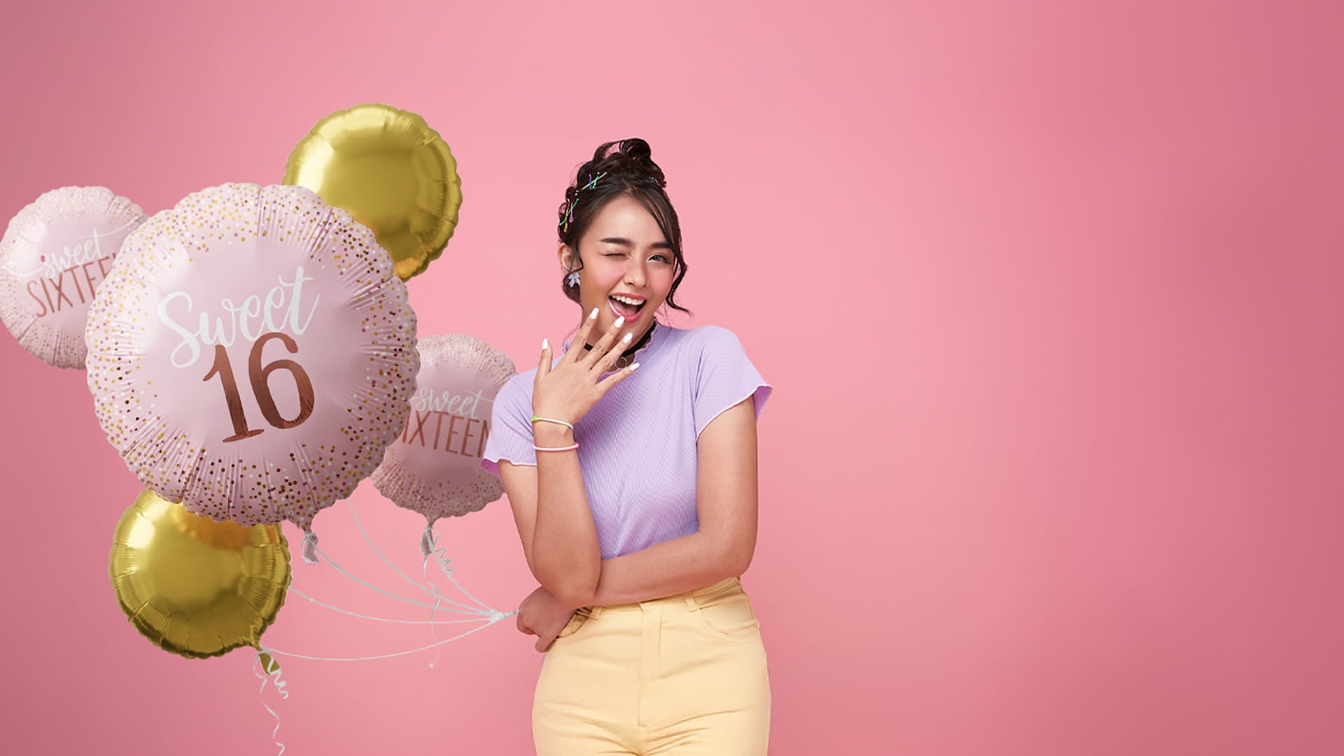 A young woman winks and smiles while holding a bunch of pink and gold "Sweet 16" foil balloons against a solid pink background.