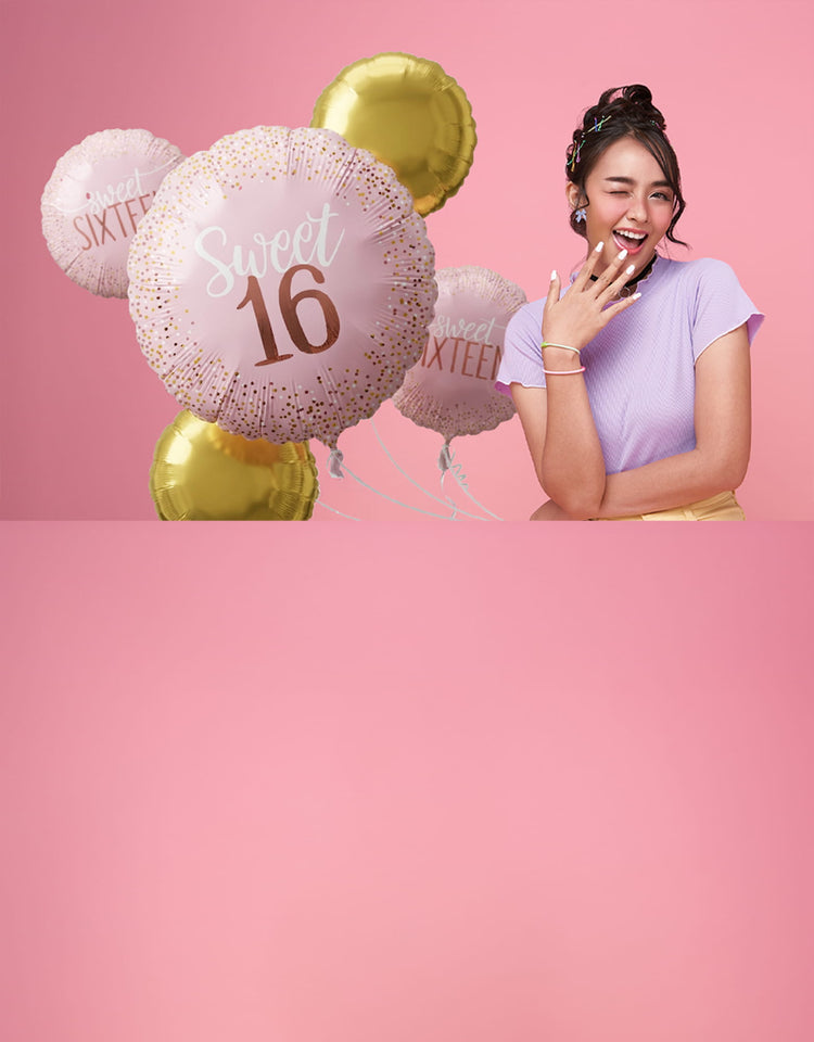 A young woman winks and smiles while holding a bunch of pink and gold "Sweet 16" foil balloons against a solid pink background.