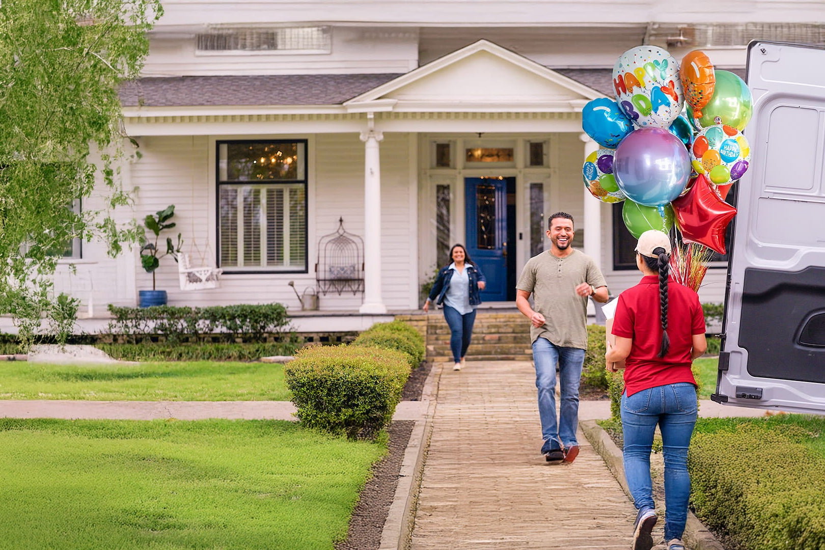 A delivery worker in a red shirt carries a large bouquet of colorful "Happy Birthday" balloons toward a smiling man and woman at the end of a suburban walkway.