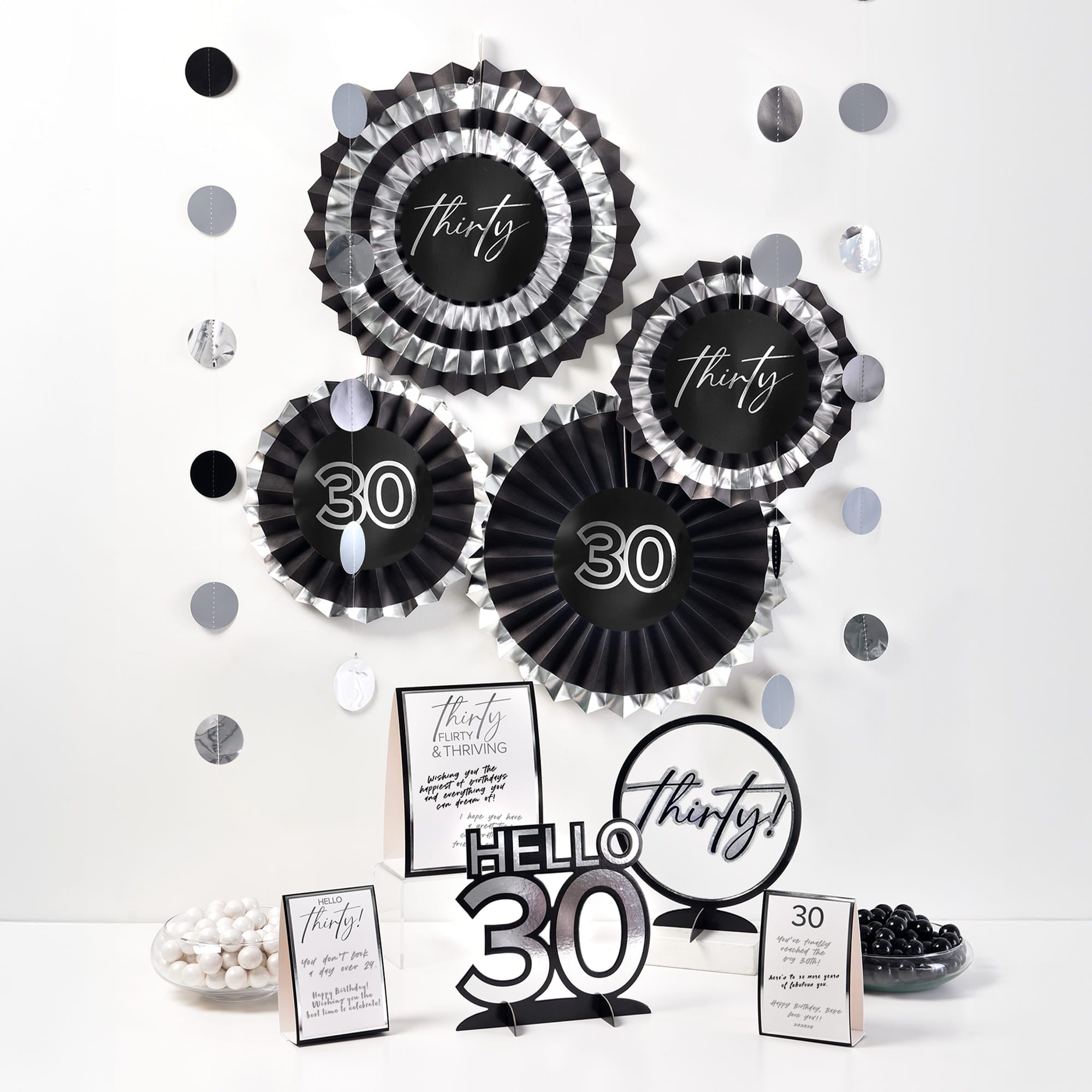 A collection of black and silver 30th birthday decorations, including hanging paper fans, a "Hello 30" centerpiece, and tabletop signs on a white surface.