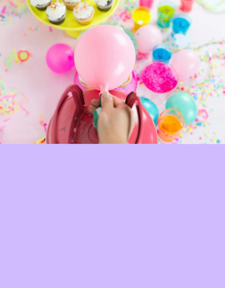 An overhead shot of a person using a pink electric pump to inflate a pink balloon amidst a colorful mess of party decorations, cupcakes, and confetti on a white surface.