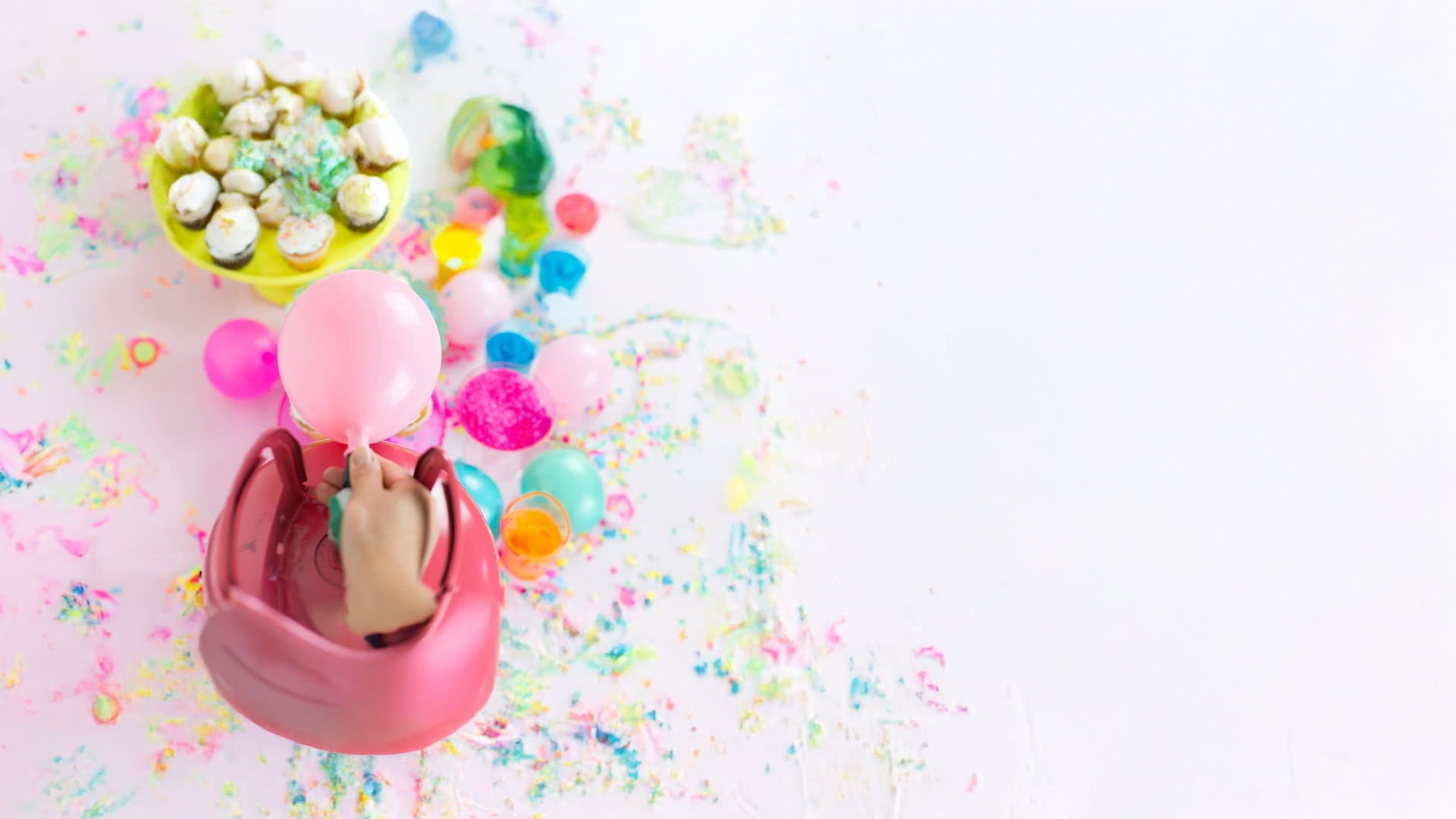 An overhead shot of a person using a pink electric pump to inflate a pink balloon amidst a colorful mess of party decorations, cupcakes, and confetti on a white surface.