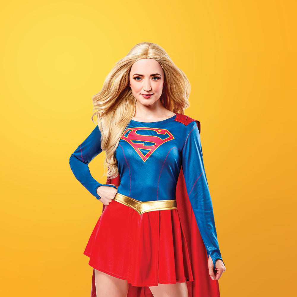 A smiling young girl in a blue and red Supergirl costume with a red cape poses against a solid yellow background.